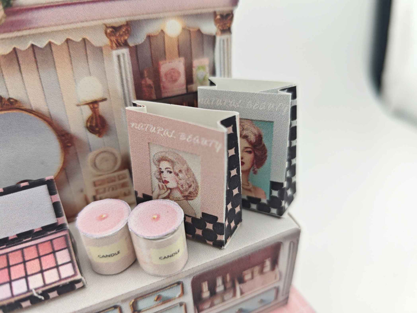 A digital image of a DIY vintage makeup counter miniature scenery kit made from cardstock. The kit features a foldable counter with a retro design, a small makeup box, a colorful poster with floral accents, and a detailed eyeshadow palette, arranged for a dollhouse display.