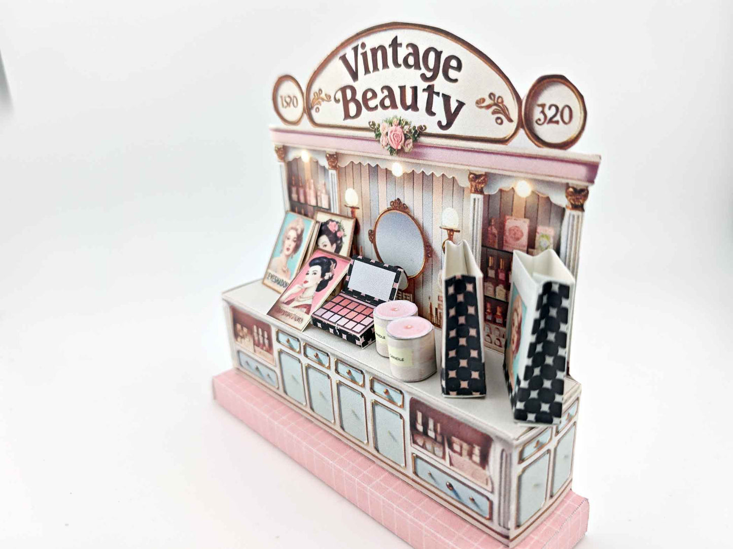 A digital image of a DIY vintage makeup counter miniature scenery kit made from cardstock. The kit features a foldable counter with a retro design, a small makeup box, a colorful poster with floral accents, and a detailed eyeshadow palette, arranged for a dollhouse display.