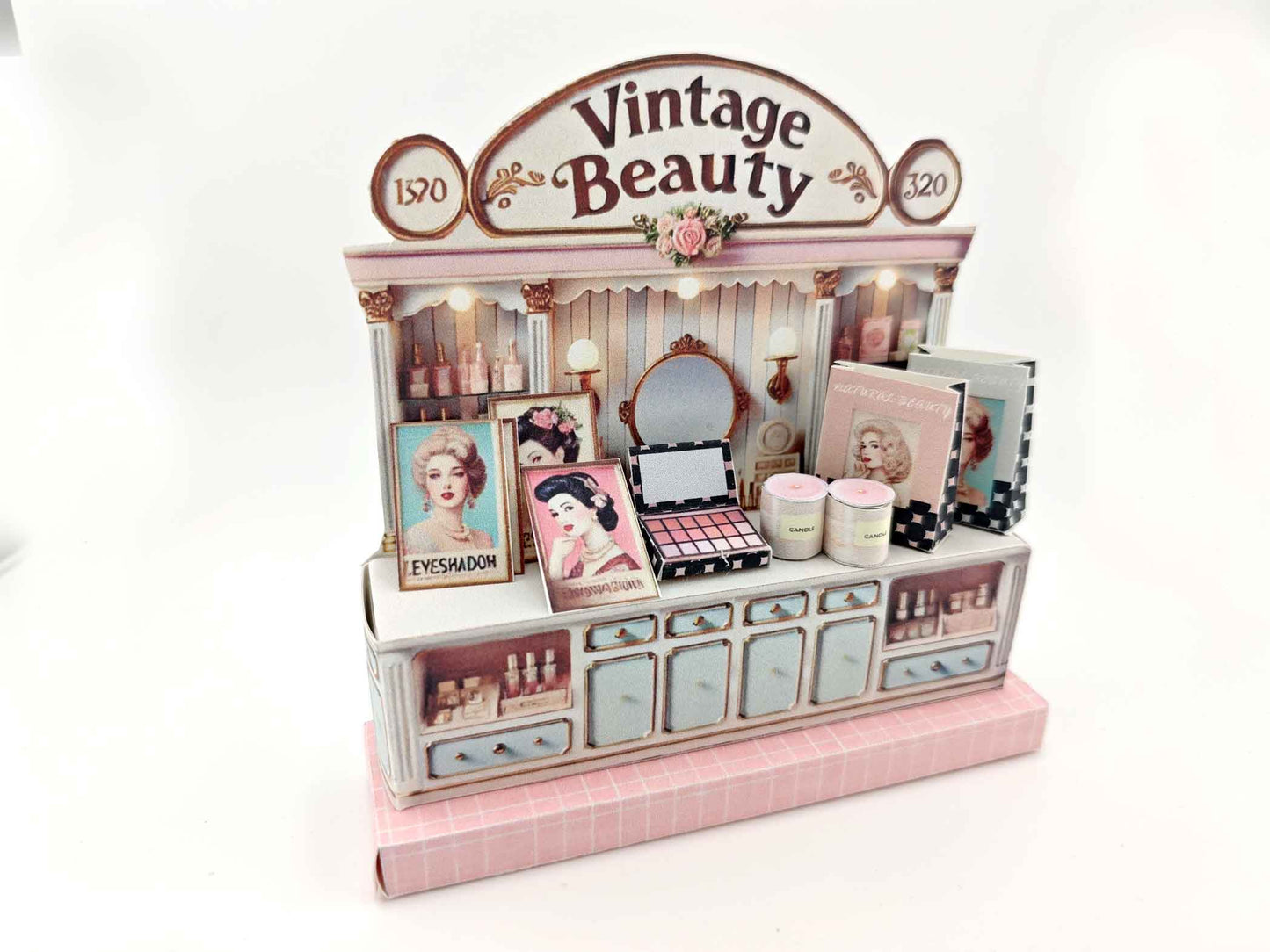A digital image of a DIY vintage makeup counter miniature scenery kit made from cardstock. The kit features a foldable counter with a retro design, a small makeup box, a colorful poster with floral accents, and a detailed eyeshadow palette, arranged for a dollhouse display.