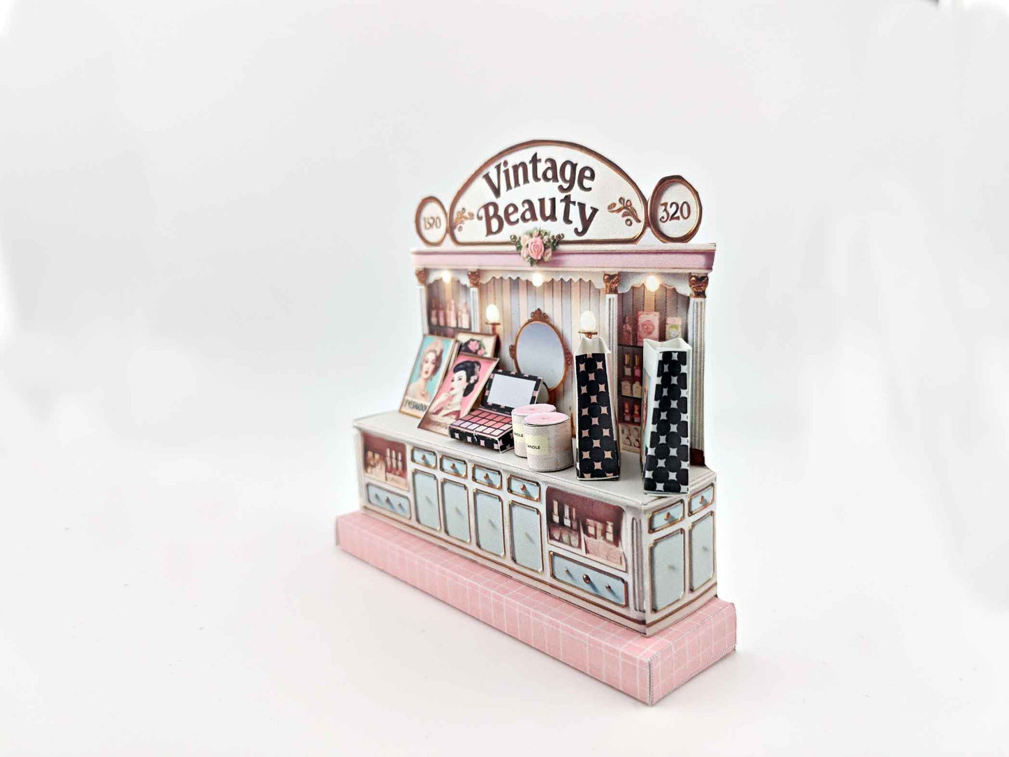 A digital image of a DIY vintage makeup counter miniature scenery kit made from cardstock. The kit features a foldable counter with a retro design, a small makeup box, a colorful poster with floral accents, and a detailed eyeshadow palette, arranged for a dollhouse display.