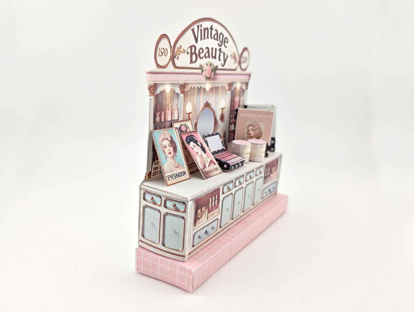 A digital image of a DIY vintage makeup counter miniature scenery kit made from cardstock. The kit features a foldable counter with a retro design, a small makeup box, a colorful poster with floral accents, and a detailed eyeshadow palette, arranged for a dollhouse display.