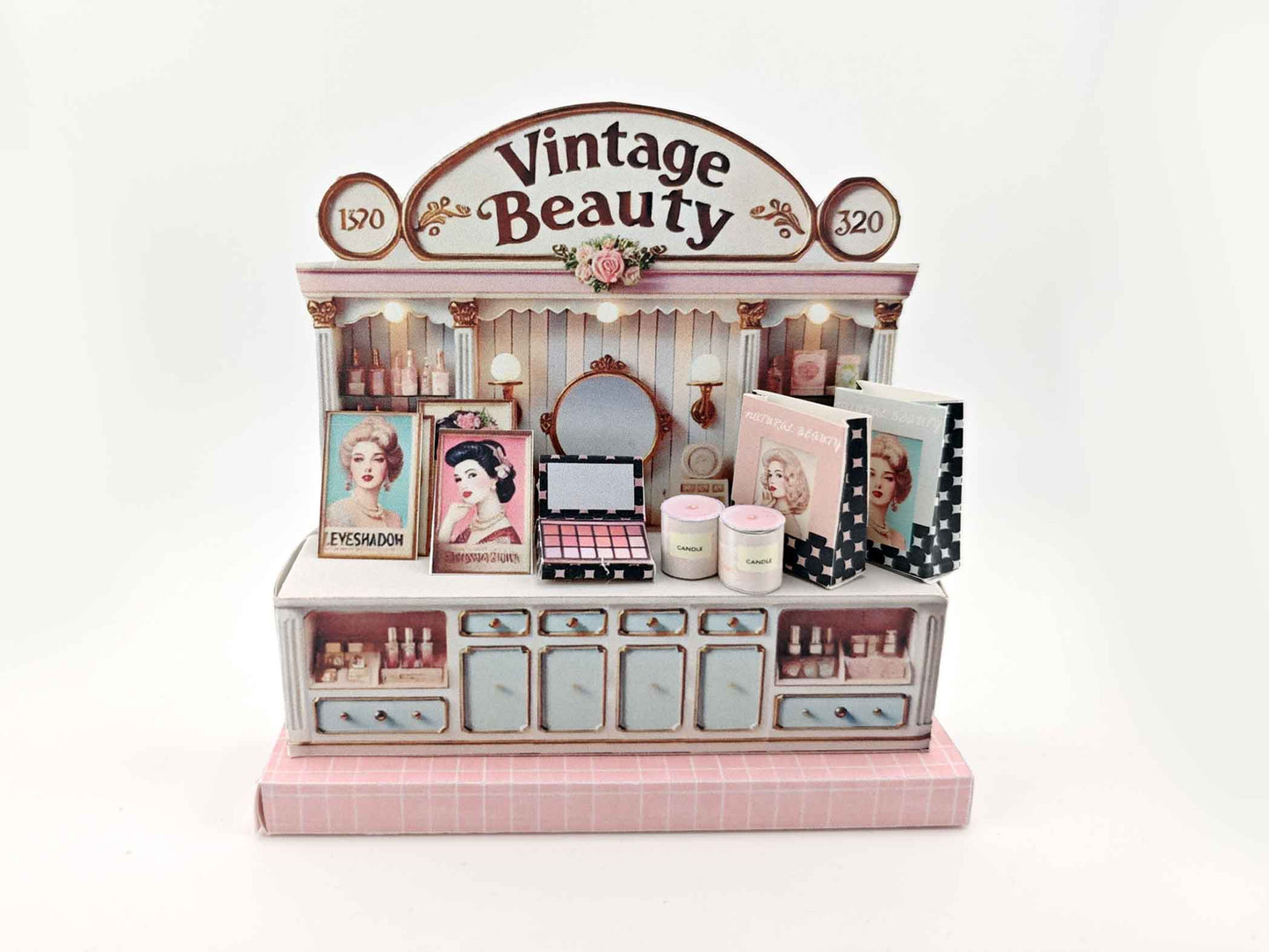 A digital image of a DIY vintage makeup counter miniature scenery kit made from cardstock. The kit features a foldable counter with a retro design, a small makeup box, a colorful poster with floral accents, and a detailed eyeshadow palette, arranged for a dollhouse display.