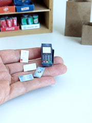 A hand holds a dark miniature credit card machine and tiny payment cards.