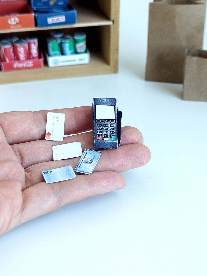 A hand holds a dark miniature credit card machine and tiny payment cards.