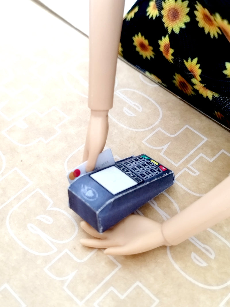 Doll's hands hold a miniature blue credit card machine with a white card inserted.