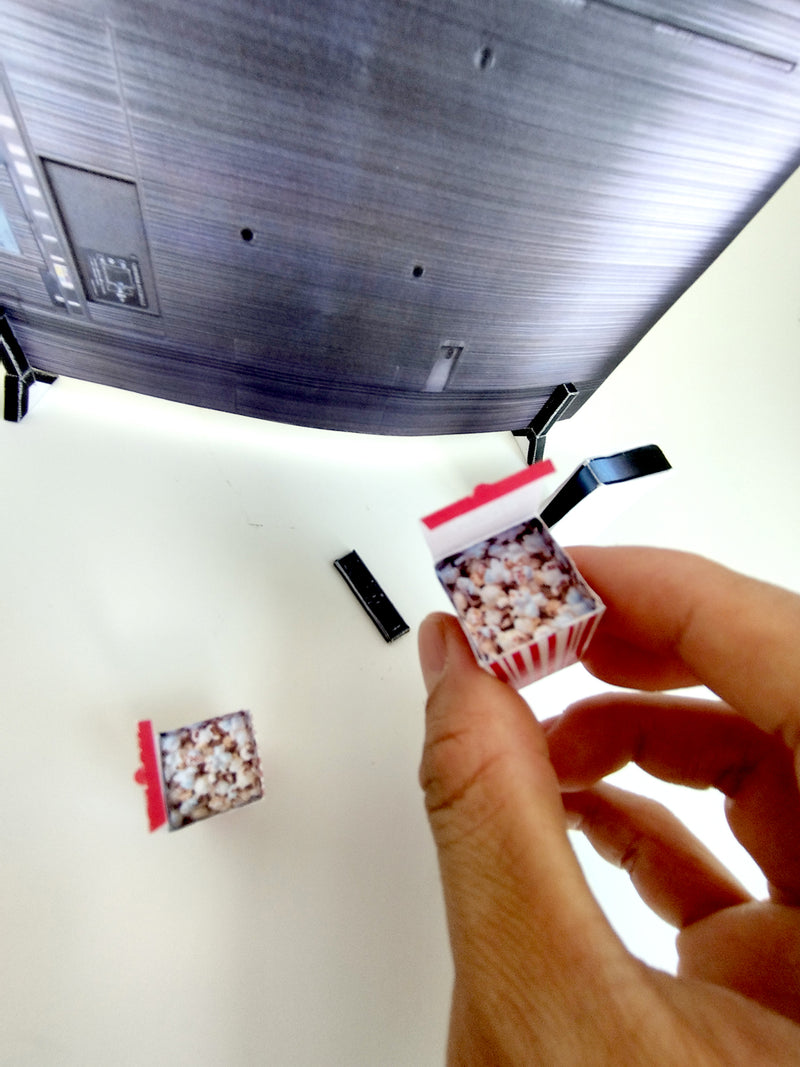 Hand holds miniature popcorn box near a curved grey TV and tiny remote.