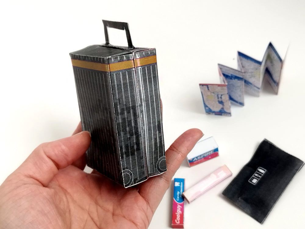 Hand holding a miniature black suitcase with gold trim and tiny travel accessories.