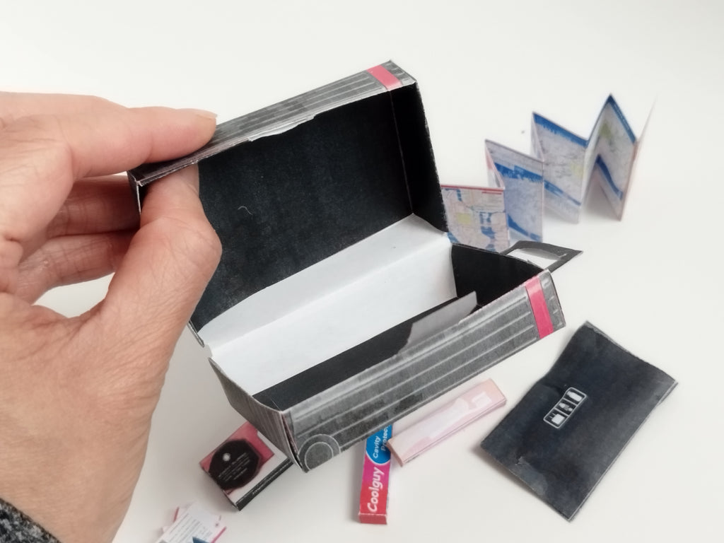 Hand holding an open miniature black and grey striped suitcase with a tiny map and accessories.