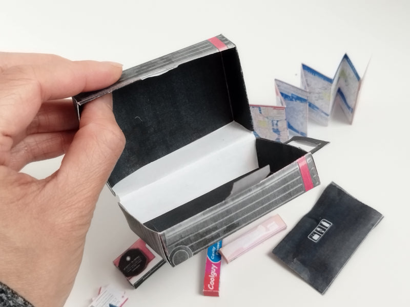 Hand holding an open miniature black and grey striped suitcase with a tiny map and accessories.