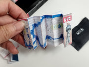 A hand holds a miniature, accordion-folded NYC map and black luggage.