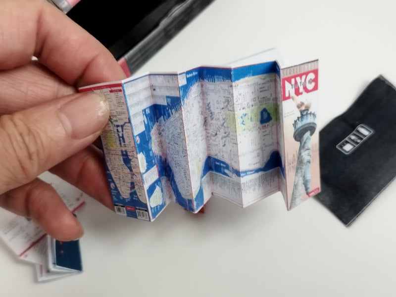 A hand holds a miniature, accordion-folded NYC map and black luggage.