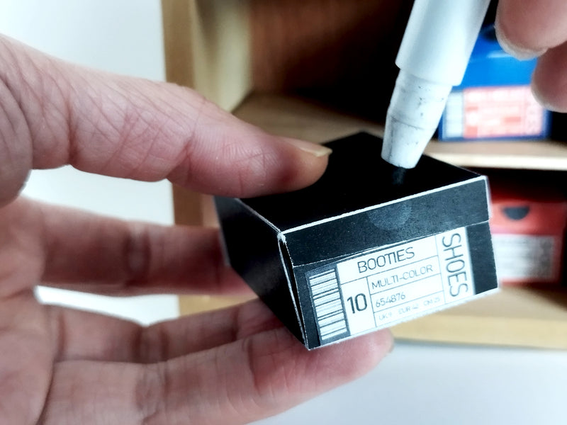 Hand holds a tiny black shoe box with a "BOOTIES SHOES" label, a marker touches it.