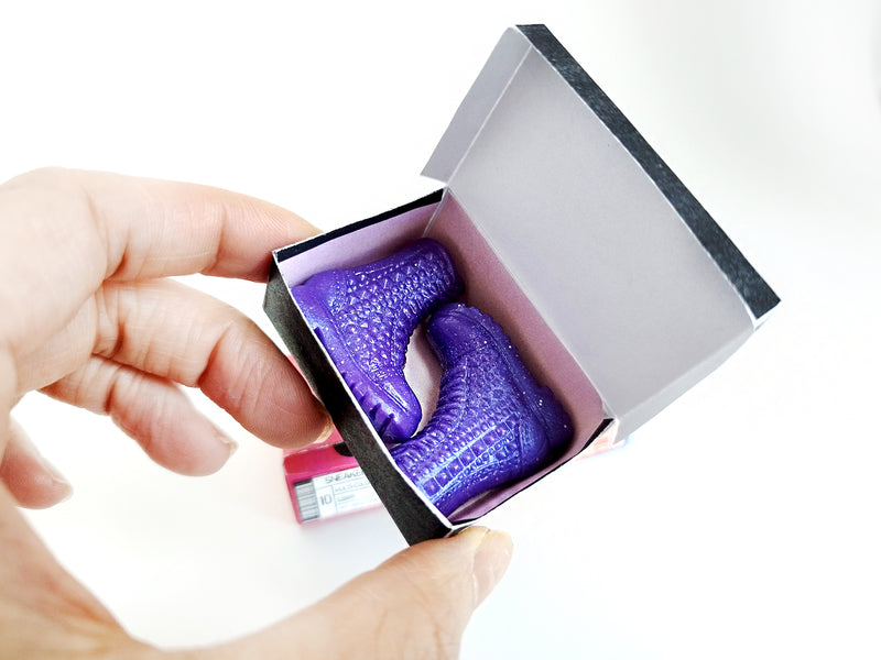 Hands hold an open black miniature box containing two shiny purple studded boots.