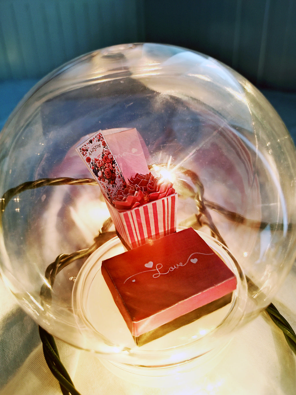 Glass dome containing miniature red flowers, a 'Love' gift box, and warm fairy lights.