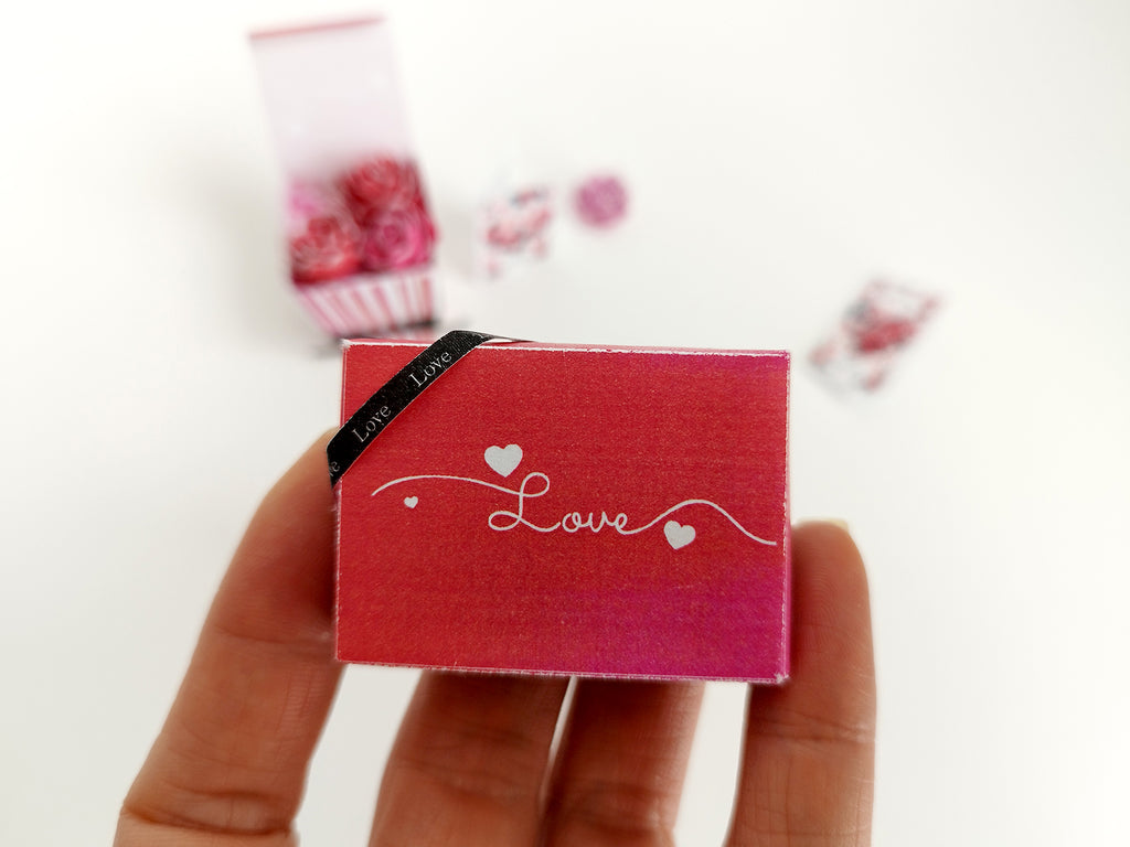 Hand holding a miniature red 'Love' gift box with a black ribbon.