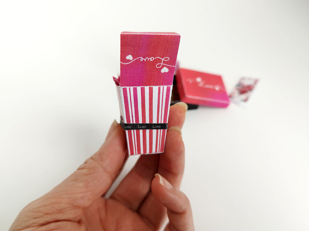 Hand holding a miniature red and white striped "Love" box with a blurred background.