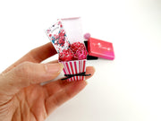 Hand holding a miniature red and white striped pot with paper roses and a card.