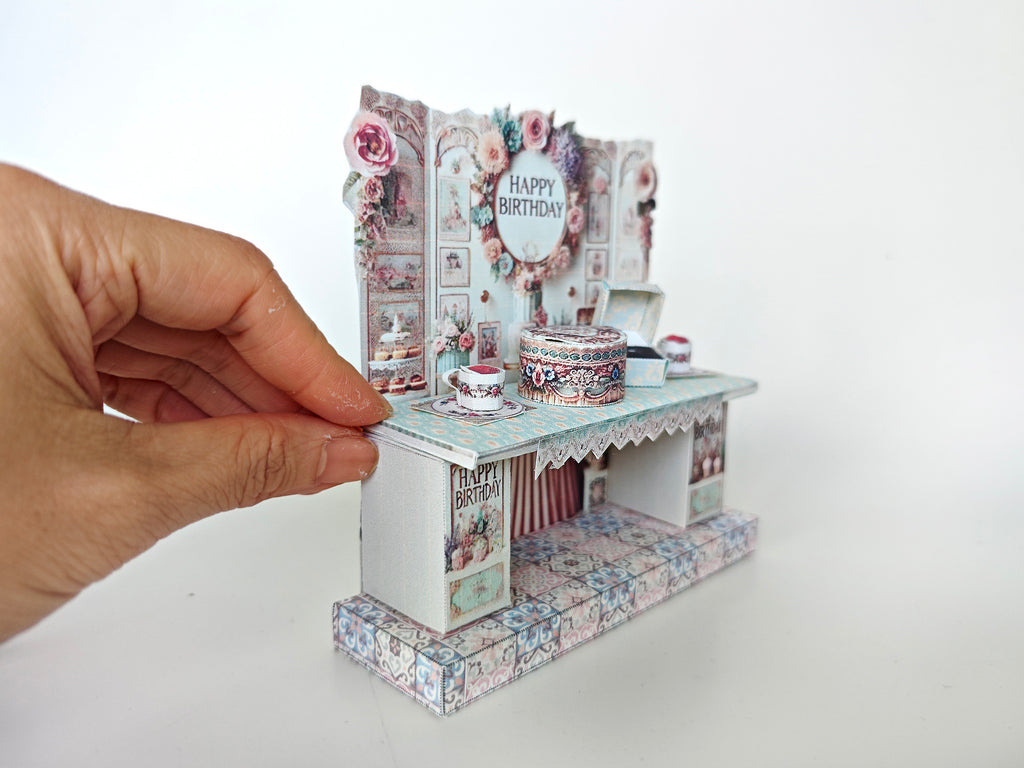 A 3D-printable STL file of a 1:12 scale vintage birthday stand DIY kit, featuring a detailed stand for displaying miniature birthday presents and cards in a dollhouse.