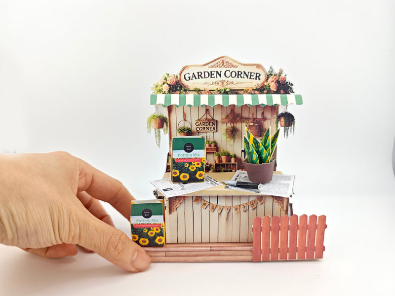 Discover the Garden Corner Miniature Stand: Create your own mini garden with plants, newspaper, potting mix, and pots. This miniature kit includes paper crafting supplies and a 7x5 frame for a charming, DIY display.