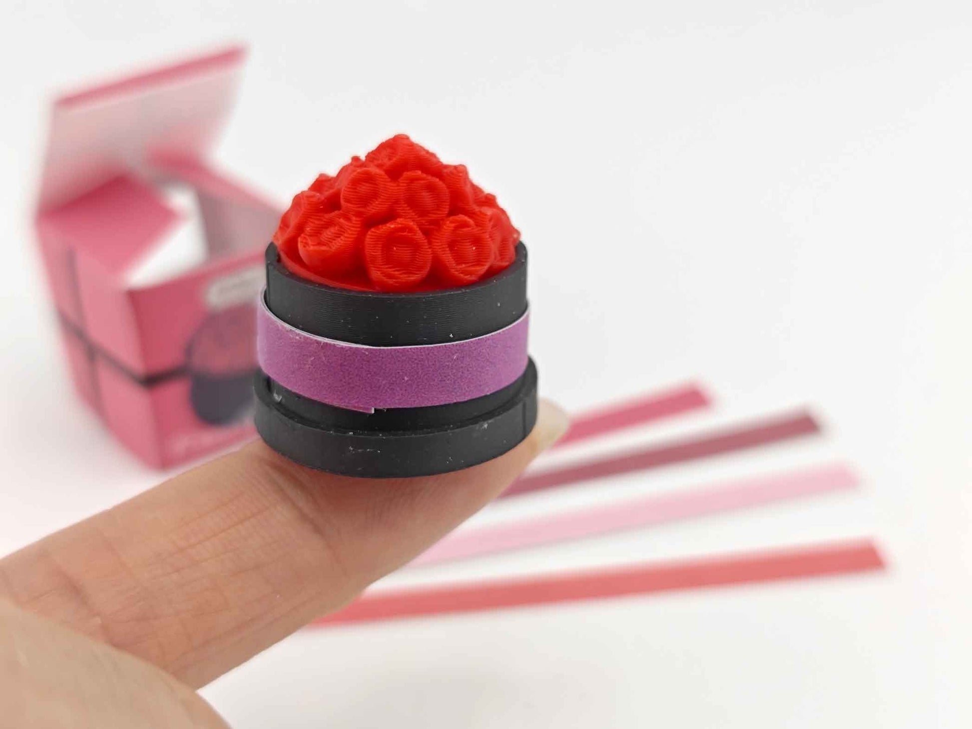 A digital rendering of a 3D printable miniature dollhouse flower box, featuring a rectangular wooden box with a grid pattern on the front. The box is accompanied by colorful strips in red, pink, and white, designed to slide into the grid for a customizable look, ideal for Mother's Day and Valentine's Day decor.