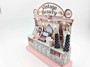 A digital image of a DIY vintage makeup counter miniature scenery kit made from cardstock. The kit features a foldable counter with a retro design, a small makeup box, a colorful poster with floral accents, and a detailed eyeshadow palette, arranged for a dollhouse display.