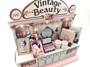 A digital image of a DIY vintage makeup counter miniature scenery kit made from cardstock. The kit features a foldable counter with a retro design, a small makeup box, a colorful poster with floral accents, and a detailed eyeshadow palette, arranged for a dollhouse display.