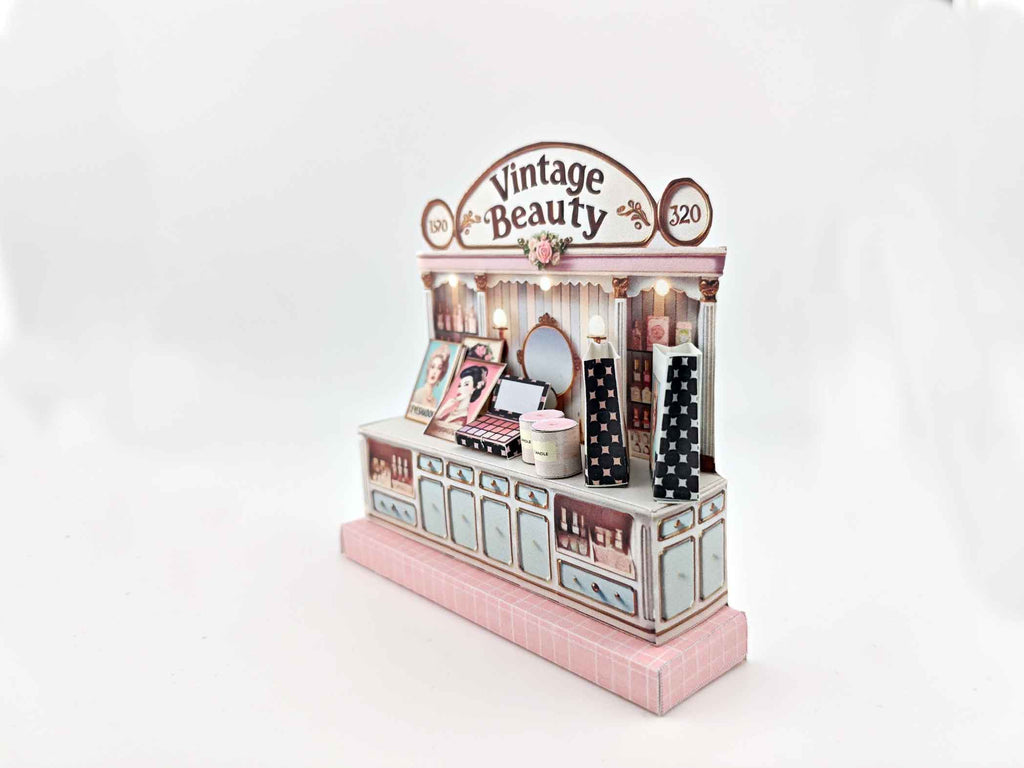 A digital image of a DIY vintage makeup counter miniature scenery kit made from cardstock. The kit features a foldable counter with a retro design, a small makeup box, a colorful poster with floral accents, and a detailed eyeshadow palette, arranged for a dollhouse display.