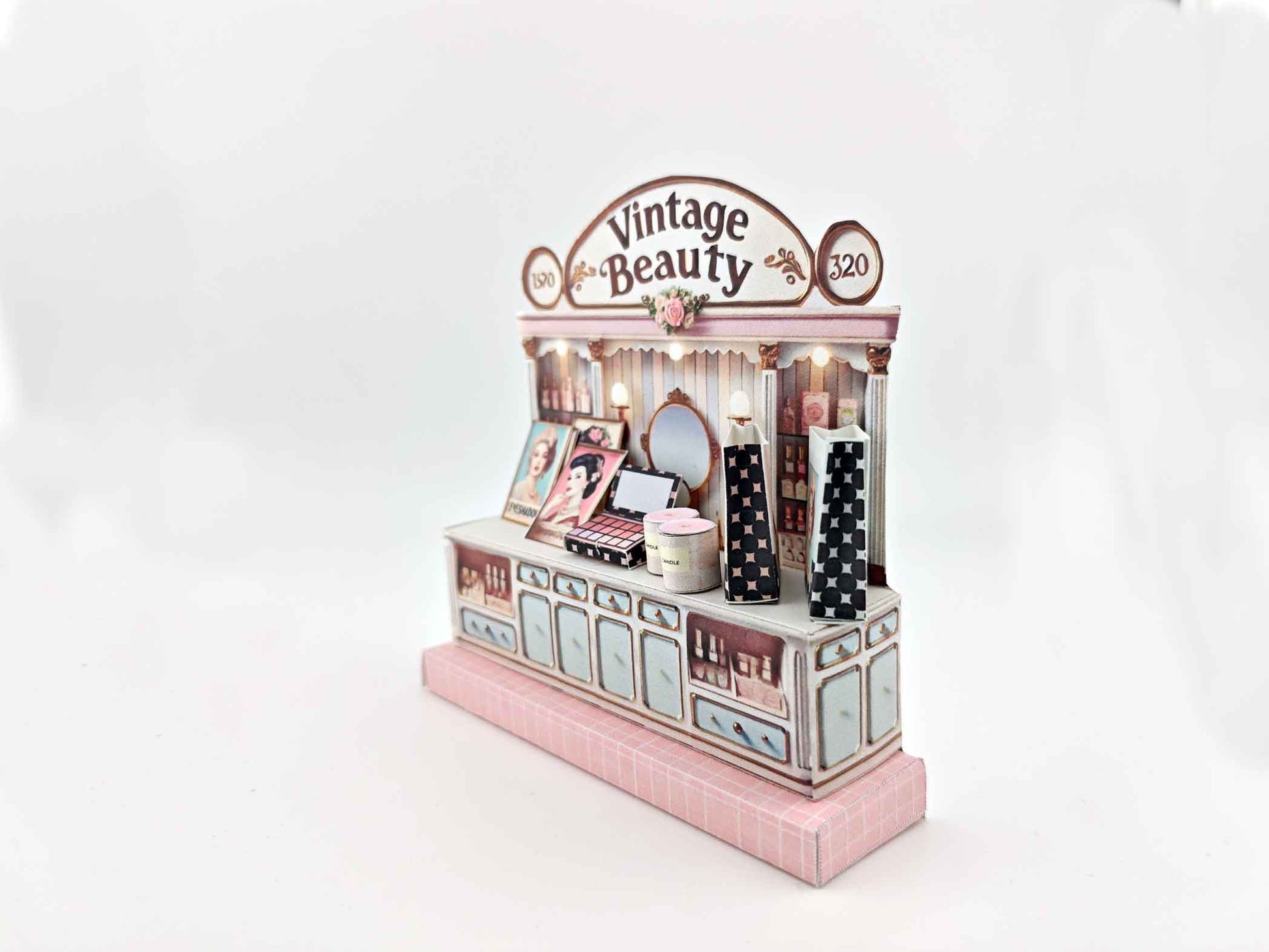A digital image of a DIY vintage makeup counter miniature scenery kit made from cardstock. The kit features a foldable counter with a retro design, a small makeup box, a colorful poster with floral accents, and a detailed eyeshadow palette, arranged for a dollhouse display.