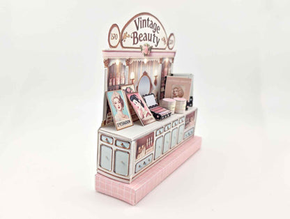 A digital image of a DIY vintage makeup counter miniature scenery kit made from cardstock. The kit features a foldable counter with a retro design, a small makeup box, a colorful poster with floral accents, and a detailed eyeshadow palette, arranged for a dollhouse display.