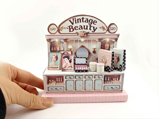 A digital image of a DIY vintage makeup counter miniature scenery kit made from cardstock. The kit features a foldable counter with a retro design, a small makeup box, a colorful poster with floral accents, and a detailed eyeshadow palette, arranged for a dollhouse display.