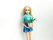 Blonde doll in blue clothes holding a miniature green plant in a pink pot.