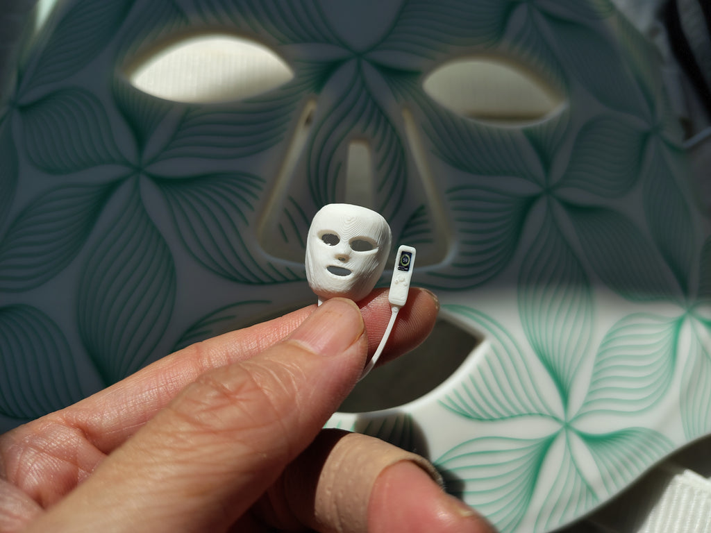 May include: A miniature white 3D-printed face mask replica with a small attached controller, held in front of a larger, full-sized white LED light therapy face mask featuring a repeating green leaf-like pattern across its surface.