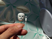 May include: A miniature white plastic facial mask with cutouts for eyes and mouth, attached by a thin cord to a small rectangular white remote control featuring a single black button, presented as a tiny replica of a light therapy device.