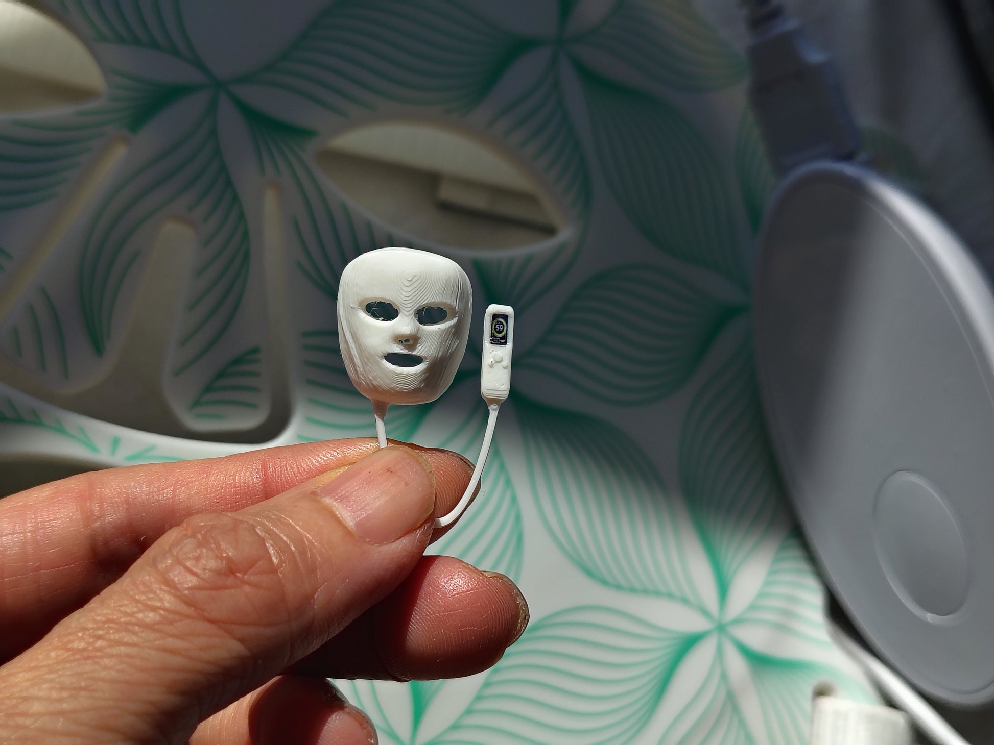 May include: A miniature white plastic facial mask with cutouts for eyes and mouth, attached by a thin cord to a small rectangular white remote control featuring a single black button, presented as a tiny replica of a light therapy device.