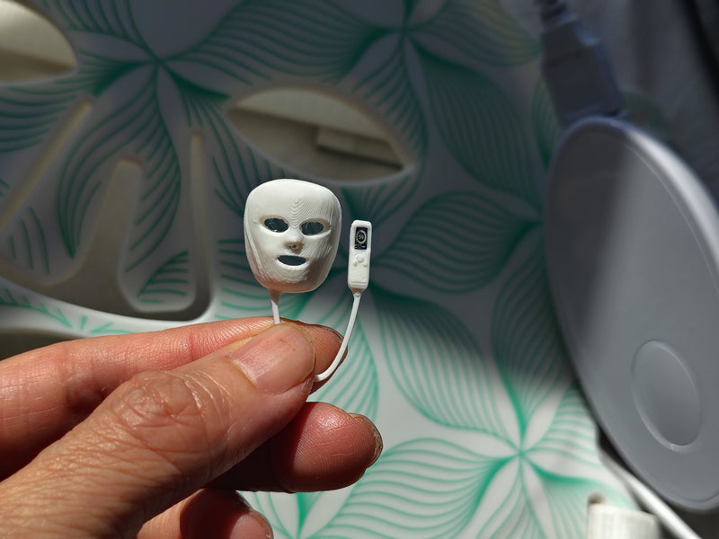 May include: A miniature white plastic facial mask with cutouts for eyes and mouth, attached by a thin cord to a small rectangular white remote control featuring a single black button, presented as a tiny replica of a light therapy device.