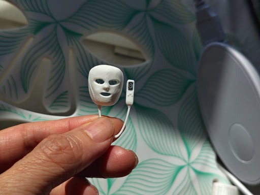 May include: A miniature white plastic facial mask with cutouts for eyes and mouth, attached by a thin cord to a small rectangular white remote control featuring a single black button, presented as a tiny replica of a light therapy device.
