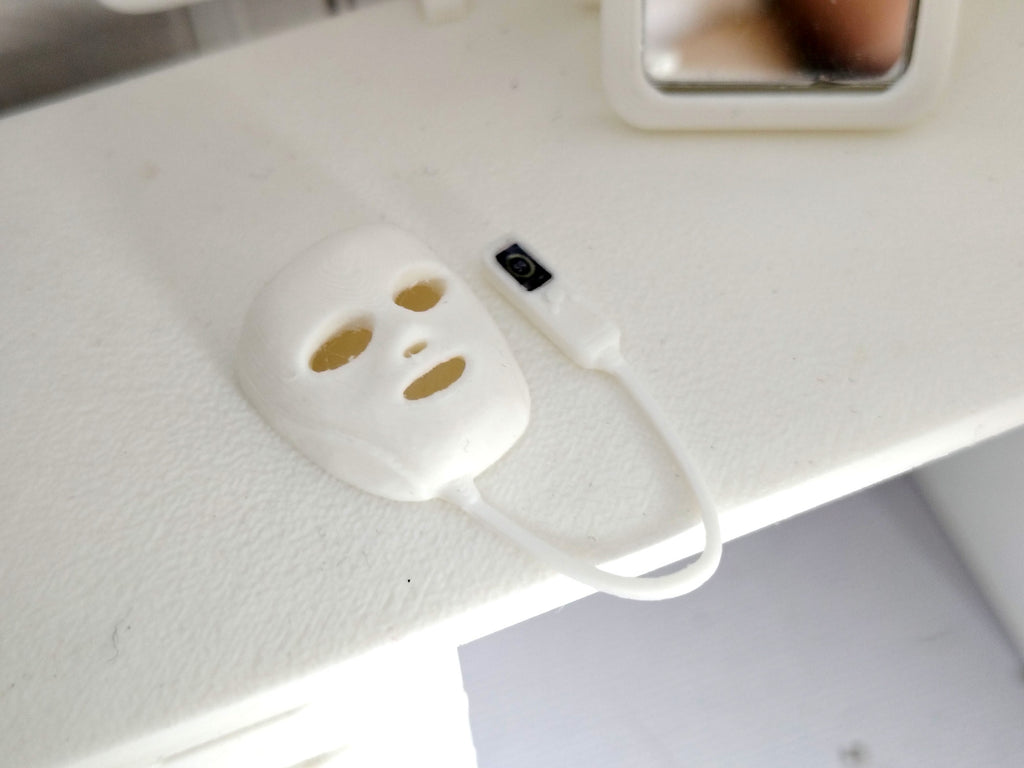 May include: A miniature white 3D printed facial mask with cutouts for eyes and mouth, connected by a thin white cord to a small white handheld remote control device featuring a black rectangular screen and small buttons on its surface.