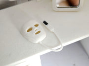 May include: A miniature white 3D printed facial mask with cutouts for eyes and mouth, connected by a thin white cord to a small white handheld remote control device featuring a black rectangular screen and small buttons on its surface.