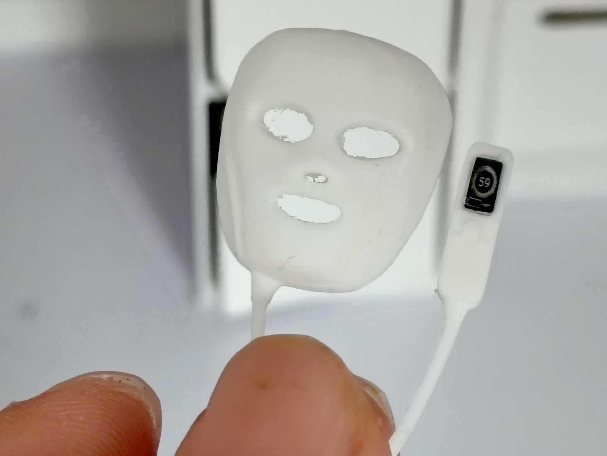 May include: A miniature white plastic facial mask with cutouts for eyes, nose, and mouth, connected by a thin white cord to a small rectangular remote control featuring a black screen with the white number 59 displayed in a circular icon.