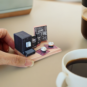 A 1:12 scale printable dollhouse Home Coffee Bar set with nine items, including a coffee machine, coffee bags, coffee cup, and desk tools, arranged for a miniature office or kitchen coffee bar diorama.