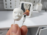 May include: A miniature white plastic facial mask with cutouts for eyes, nose, and mouth, connected by a thin white cord to a small rectangular remote control featuring a black screen with the white number 59 displayed in a circular icon.